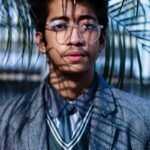Portrait of a young man wearing eyeglasses and a suit in a natural setting with palm leaves.
