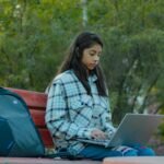 South Asian woman in plaid shirt using laptop on bench in park setting.